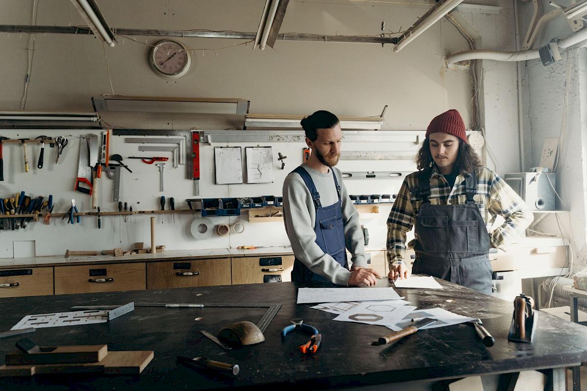 Zwei Handwerker besprechen Zeichnungen an der Werkbank einer hellen Werkstatt